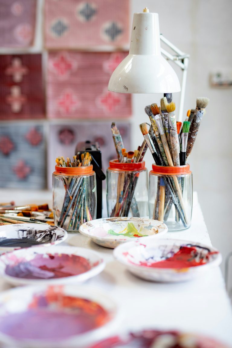 three glass jars with paint brushes on a table and plates with paint
